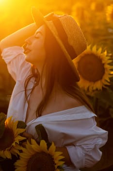 A girl in a hat on a beautiful field of sunflowers against the sky in the evening light of a summer sunset. Sunbeams through the flower field. Natural background