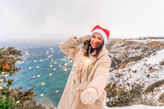 Outdoor winter portrait of happy smiling woman, light faux fur coat holding heart sparkler, posing against sea and snow background.