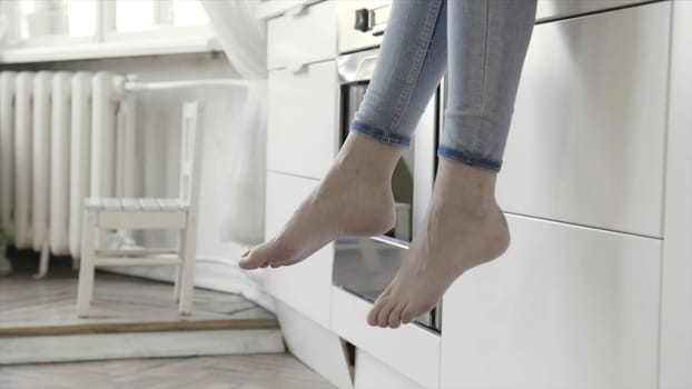 Close up for woman legs dangling with white kitchen drawers and oven on the background. Women wearing jeans sitting barefoot on kitchen counter.