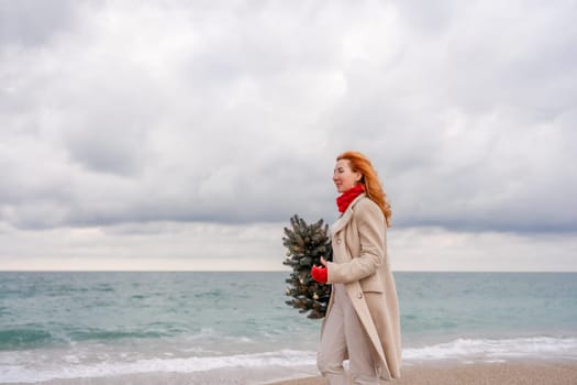 Redhead woman Christmas tree sea. Christmas portrait of a happy redhead woman walking along the beach and holding a Christmas tree in her hands. Dressed in a light coat, white suit and red mittens