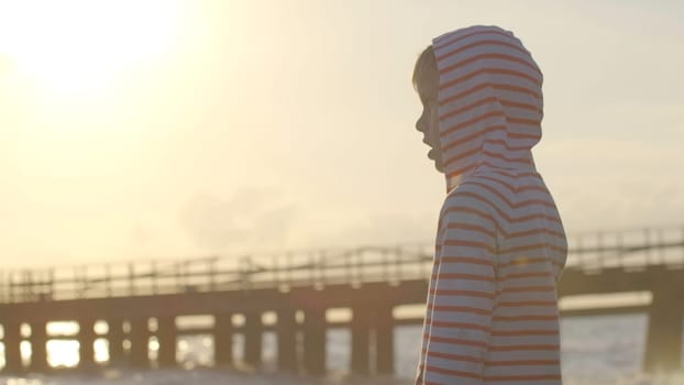 Boy looks at sea on background of pier. Creative. Boy looks at horizon during sunset on beach. Child looks in amazement at sea at sunset in summer.