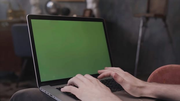Over the shoulder view of a man sitting at his desk and working on a laptop with Green Screen. Man using notebook computer with chroma key.