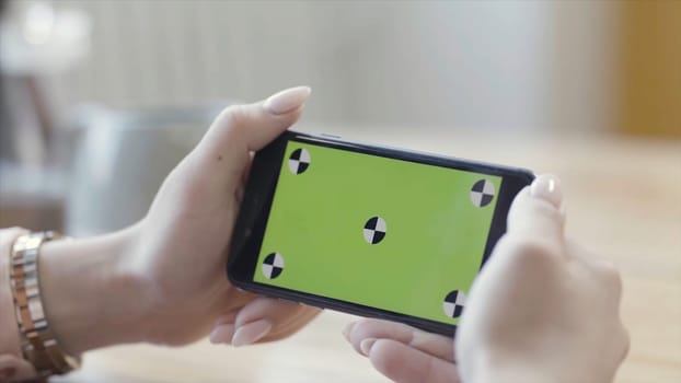 Woman looking at horizontal smartphone with green screen. Close up of beautiful female hands holding mobile phone with chromakey on blurred room background.