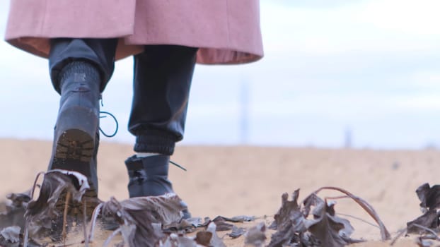 Rear view of female black shoes on the sand, close up view. Woman wearing black leather boots and pink coat walking away on sandy coast with dry tree leaves.