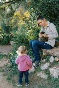 Little girl looks at her mother stroking a cat in her arms while sitting on a park bench. Back view. High quality photo