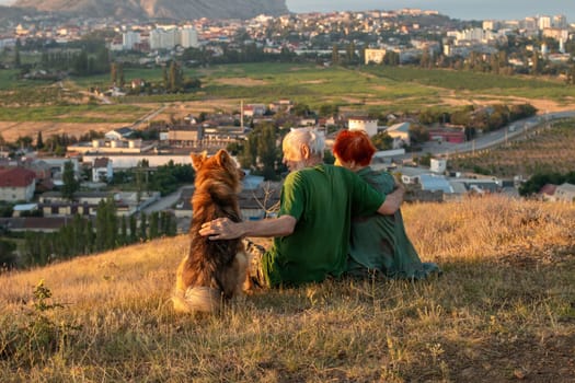 An elderly couple sits on a mountain with their backs with a beautiful view of the mountains and the sea in the distance, a dog sits next to them.