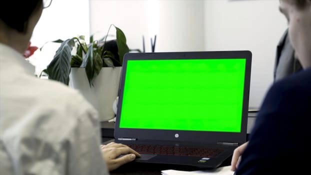 Young women working in office, sitting in chairs in front of computer with green screen, rear view. Two girls co workers looking at computer monitor with chroma key over white wall background
