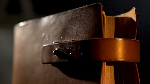 Vintage leather book with yellow pages. Close up of an ancient notebook with brown leather cover and a strap in vertical position isolated on black background under the dim light.
