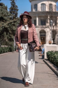 Woman park city. Stylish woman in a hat walks in a park in the city. Dressed in white corset trousers and a pink jacket with a bag in her hands