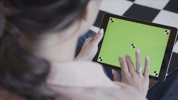 Young woman using black tablet device with green screen. Woman holding tablet, zooming pages while sitting on the couch in the living room. Chroma key.