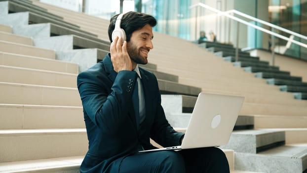 Professional business man sitting at stairs while working on laptop. Skilled project manager listening music from headphone and checking email and discussion about marketing plan. Outdoor. Exultant.