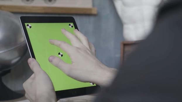 Hands of an unknown young guy tapping and scrolling on laptop with green screen on wooden table next to silver globe. Man using his Ipad with chroma key.