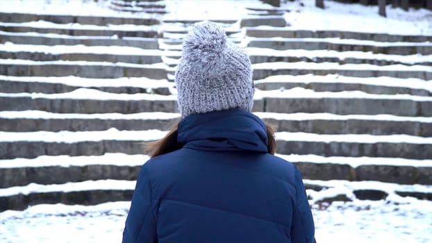 Rear view on woman with hat stays in a park while snowing, snowy stairs background. Alone woman stand alone. snow in winter, landscape, romantic scene