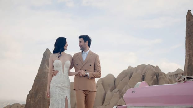 An interesting beautiful photo shoot of the newlyweds. Action. The bride of pretty appearance in a narrow beautiful white long dress with her cute groom in a brown suit are photographed posing next to a pink car hugging and looking at each other