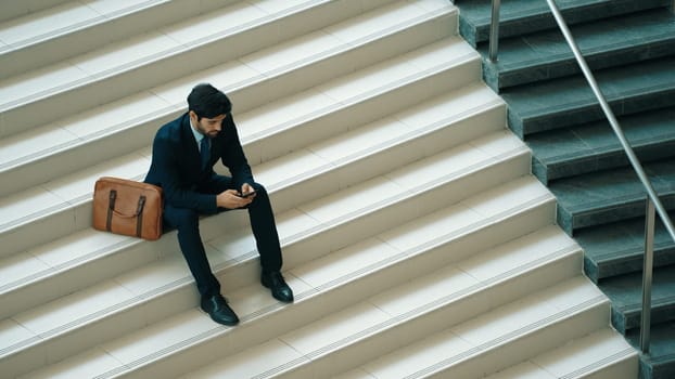 Business man sitting at stair while using mobile phone plan business strategy. Skilled project manager working by using smart phone contact business team or investor while wearing suit. Exultant.