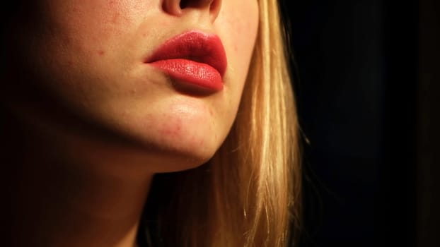 Close-up of woman with blond hair applying the fashion red lipstick on the dark background. Beauty concept