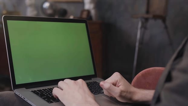 Over the shoulder view of a man sitting at his desk and working on a laptop with Green Screen. Man using notebook computer with chroma key.