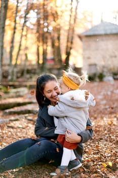 Little girl hugs her smiling mother sitting on the ground in the autumn forest under falling leaves. High quality photo