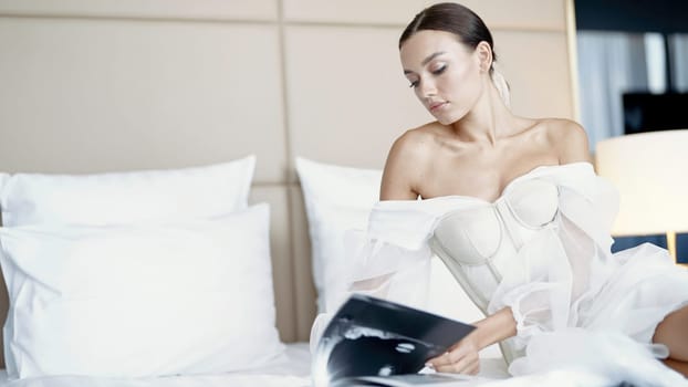 A young well-groomed model in a white dress on the bed. Action. A woman carefully leafing through a magazine lying in a bright spacious room. High quality 4k footage