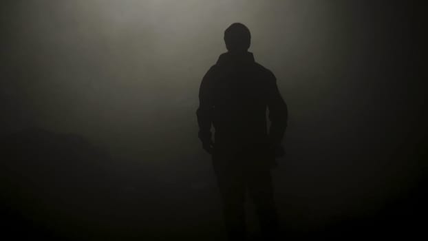 Black silhouette of man standing in smoky dark. Stock footage. Mysterious silhouette of young man stands in darkness lit only by dim light in haze.