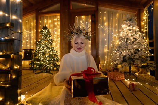 Happy blonde as Snow Maiden in white costume. Wooden house exterior Festive setting. Character with crown creating festive scene