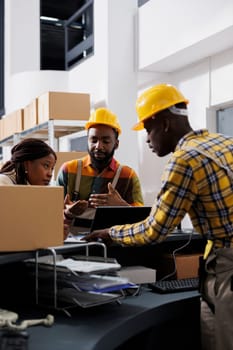 Postal service workers checking parcel invoice on laptop and clipboard. African american men and woman storehouse employees team managing inventory documentation at reception