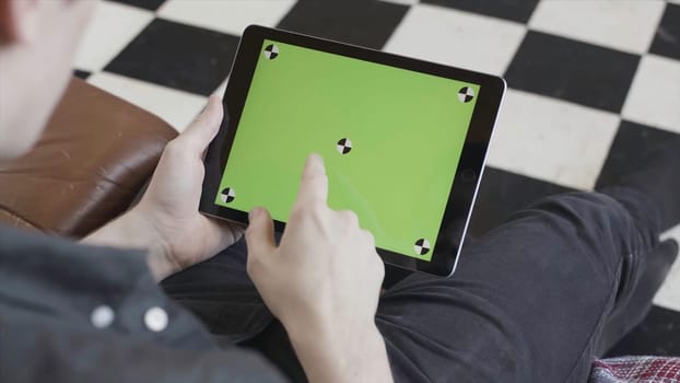 Technology and internet concept, over the shoulder view of a man tapping on the green screen of a tablet with tracking marks. Close-up of man in black shirt using his device with chroma key.