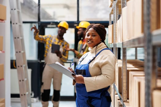 Smiling warehouse woman distribution manager looking at camera and writing on clipboard. African american storehouse manager wearing workwear overall standing in storage room portrait