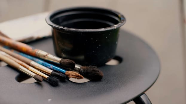 Close up of paint brushes with colorful stains and a small cup of water on a chair. Video. Concept of art and creativity