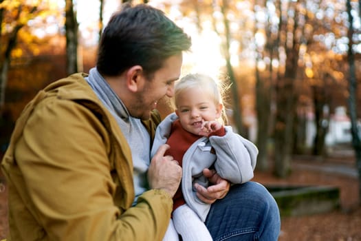 Dad hugging smiling little girl sitting with her in autumn park. High quality photo