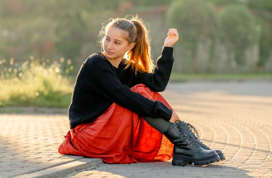 Beautiful teenager girl in red skirt sitting outdoors at street. Pretty teen model posing in trendy clothes