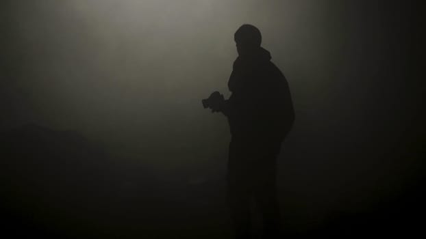 Black silhouette of man standing in smoky dark. Stock footage. Mysterious silhouette of young man stands in darkness lit only by dim light in haze.