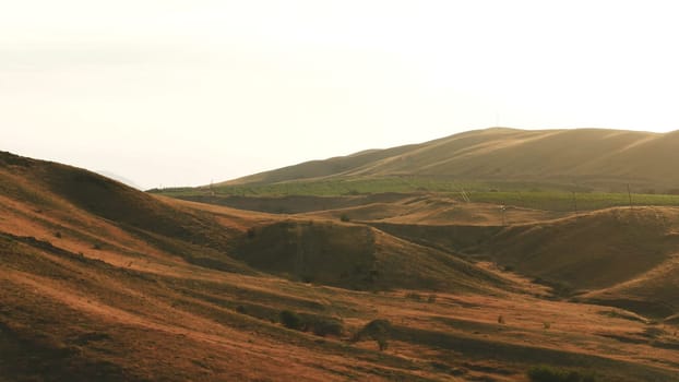 Aerial view of exciting red hills covered with shrubs and large green farmlands against the evening sky in summer day. Amazing colorful landscape
