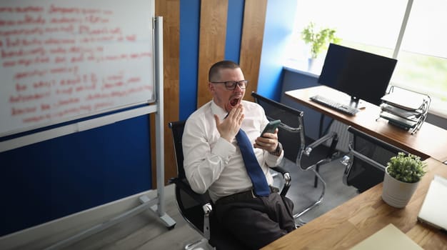 Businessman looks into smartphone and yawns at workplace. Boredom and laziness at work concept