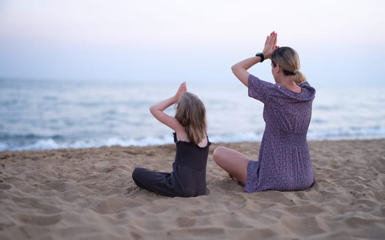 Mom and daughter doing yoga and sitting in lotus position on seashore back view. Pilates sports training with kids concept