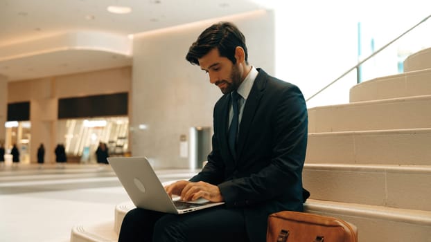 Skilled investor working or planing strategy by using laptop at stair. Professional business man wearing suit while working and typing data analysis by using laptop at modern hotel. Exultant.