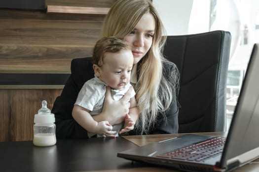 Concept of combining business and caring for newborn child, young businesswoman holds small child in arms and communicates via video link via laptop in office.