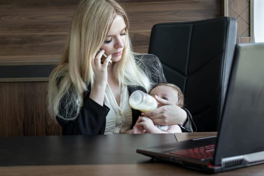 Young business woman feeds baby from bottle in office and talks on phone, concept of combining business and caring for newborn baby.