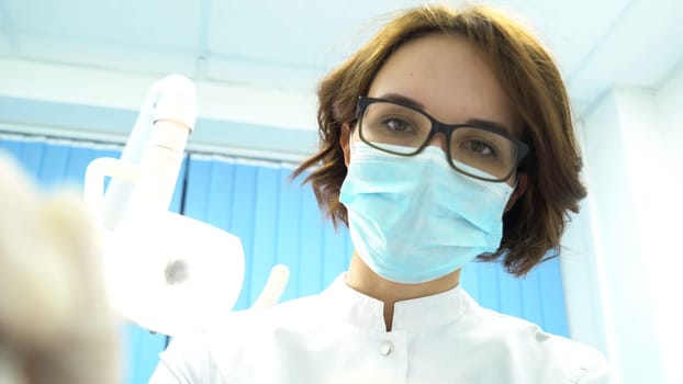 First person view of a young female dentist in mask approaches with Tools. Young doctor standing over the patient, looking at camera.