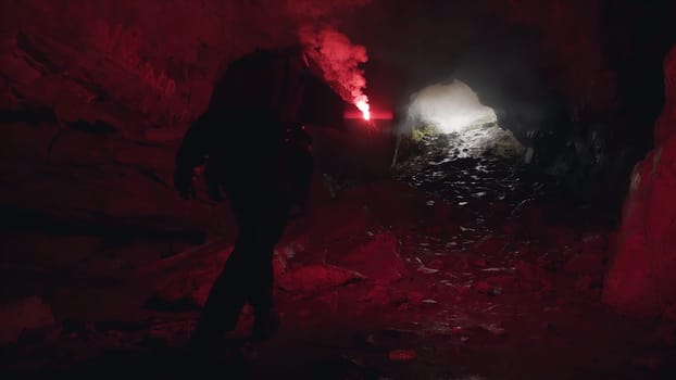 Man lighting his way inside dark cave with a burning signal flare and leaving the entrance behind him. Dangerous tourism inside dark grotto.