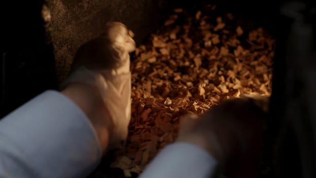 A man puts sawdust in a cast iron furnace close-up. Man's hands putting wood into the fire close up. Wood furnace. Open firebox with a hardwood log laid inside a furnace. Flames and an ember are visible in a blurry background.