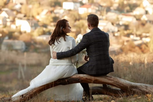 bride in white wedding dress and groom are sitting on tree in nature, back view