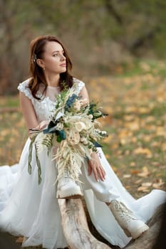 beautiful bride wearing white boots sitting on tree and holding autumn bouquet.