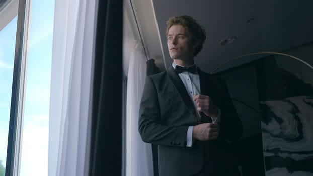 Handsome man in suit by window. Action. Gentleman in suit with bow tie looks out window. Attractive man in suit is preparing for celebration.