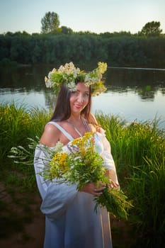 Adult mature brunette woman in a white dress, sundress and a wreath of flowers in summer by the water of river or lake in evening at sunset. Celebration of the Slavic pagan holiday of Ivan Kupala