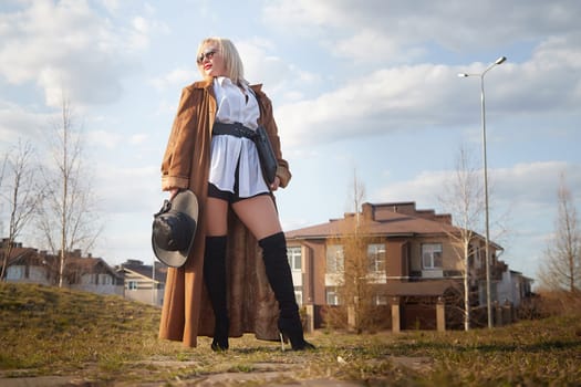 stylish, fashionable, imposing blonde girl in black glasses, a white shirt, a short skirt and long raincoat with a laptop and a hat on the street of the town. Defiantly dressed confident businesswoman