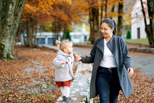 Mom looks at a little girl leading her by the hand along a stone fence in the autumn forest. High quality photo