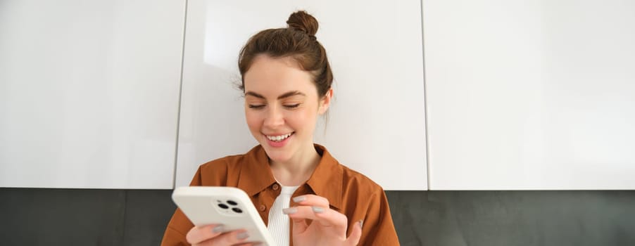 Happy girl with smartphone, looking at phone and smiling, standing at home, concept of buying online, shopping in mobile app.