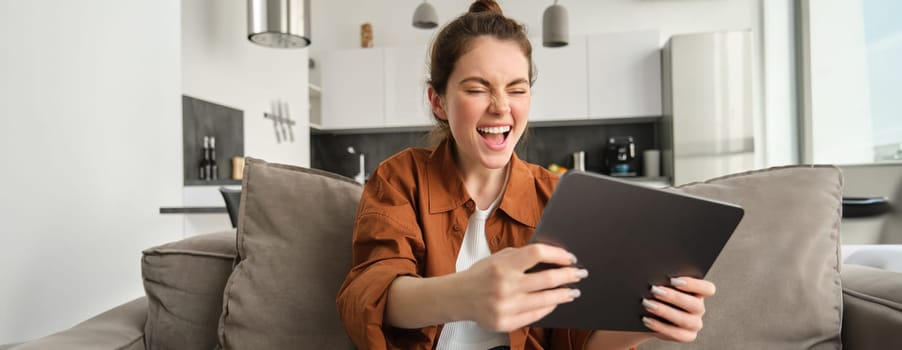 Portrait of excited young woman with digital tablet, sitting on couch, laughing and smiling, winning on video game, spending time at home.