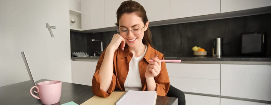 Young woman making notes, sitting in kitchen with pen and notebook, smiling while writing in her planner, planning her schedule.